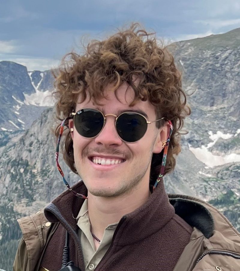 A smiling person with curly hair wearing round sunglasses and a jacket with a mountainous background.