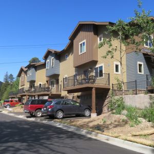 A row of modern, multi-story townhouses with garages and balconies, situated along a paved street with parked cars in front. Trees and a mountain are visible in the background.