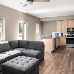 A modern, open-concept living room and kitchen with a gray sectional sofa, an island with barstools, wood cabinets, stainless steel appliances, and wood flooring. Light streams in through large windows.