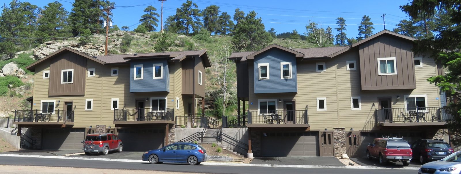 Two adjacent two-story houses with garages and balconies, situated on a sloped hillside. A red vehicle, a blue vehicle, and an SUV are parked in front. Hills with scattered trees are seen in the background.