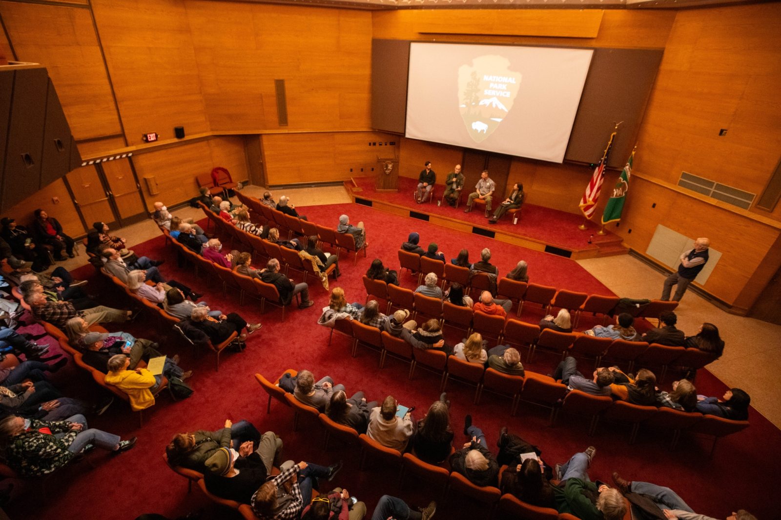 Audience seated in an auditorium watching a panel discussion, with a screen displaying "National Parks" and flags in the background.
