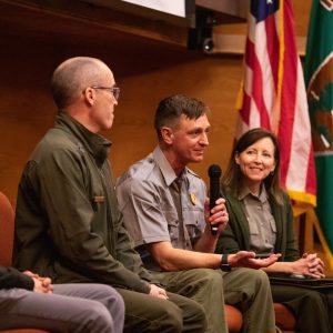Four people seated on a panel, with one person holding a microphone. U.S. and organizational flags are visible in the background.