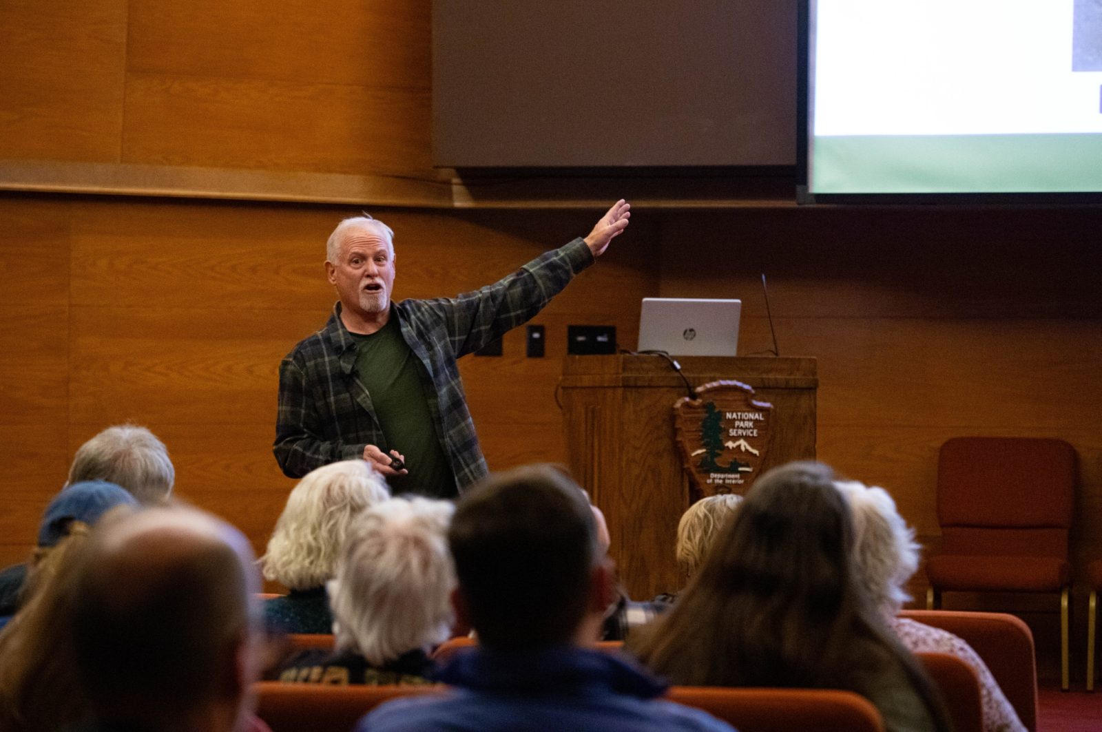 A person gestures while presenting a lecture in a wood-paneled room, with an audience seated in front.