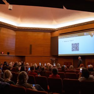 Audience in a wood-paneled lecture hall listens to a presentation with a slide displaying a QR code and text about mountains.