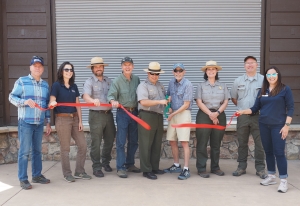 Nine people, including park rangers and civilians, stand in a row in front of a building as two people cut a red ribbon, suggesting a ribbon-cutting ceremony or event.