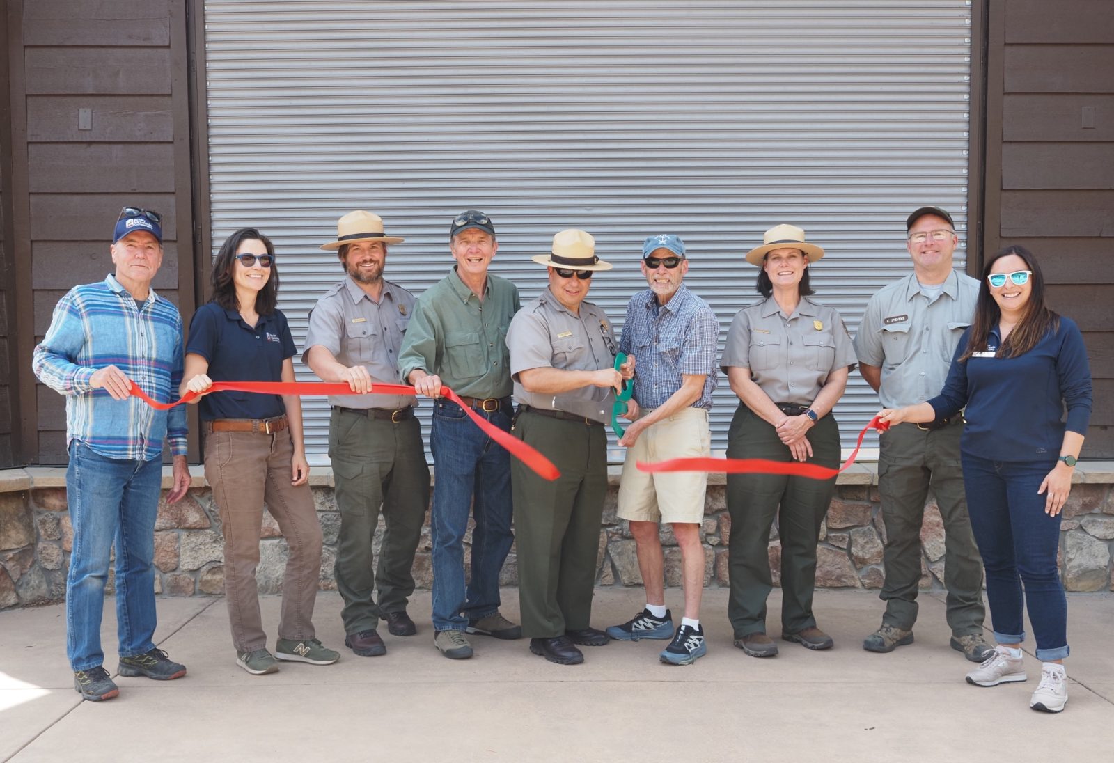 Nine people, including park rangers and civilians, stand in a row in front of a building as two people cut a red ribbon, suggesting a ribbon-cutting ceremony or event.
