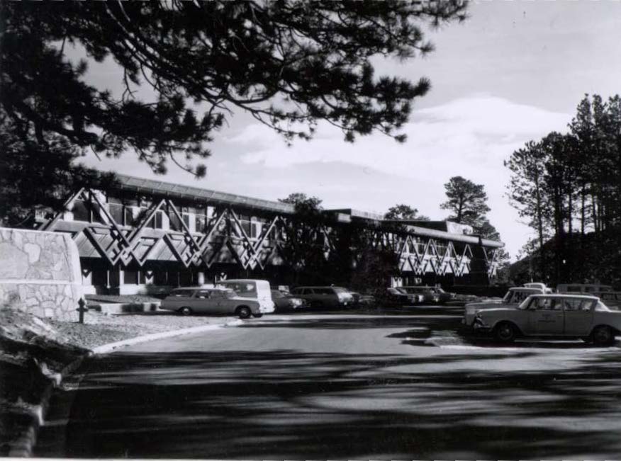 Black and white photo of a mid-century building with geometric facade, several parked cars, trees, and a stone wall in the foreground.
