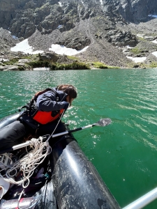Person wearing a life jacket leans over the edge of an inflatable boat with a paddle, on a green mountain lake surrounded by rocky terrain and patches of snow.