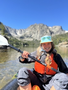 A person wearing a life jacket and holding a paddle sits in a kayak on a lake, with mountains and trees visible in the background under a clear blue sky.