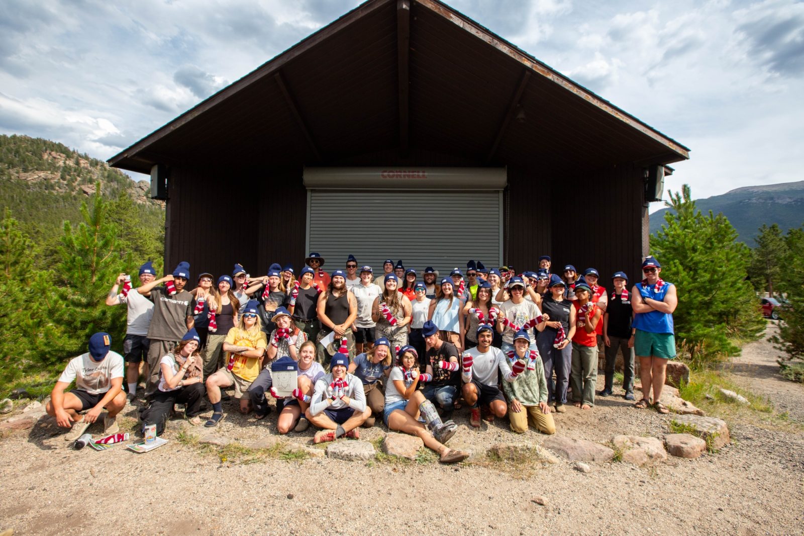 A large group of people pose together outdoors in front of a cabin, surrounded by trees and mountains, on a sunny day.