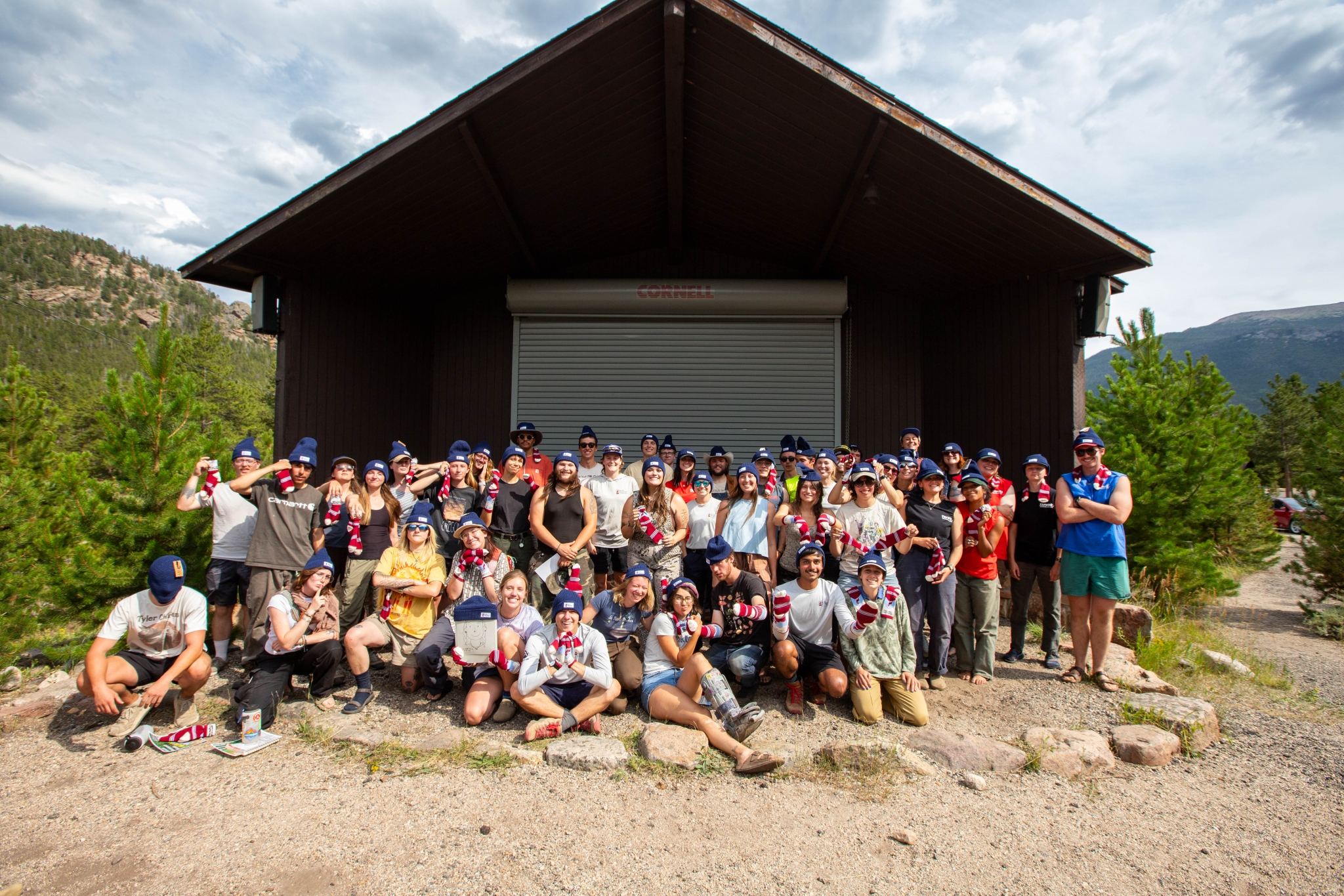A large group of people pose together outdoors in front of a cabin, surrounded by trees and mountains, on a sunny day.