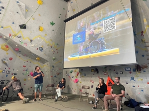 A man speaks into a microphone beside a projected slide showing a person in a wheelchair on a paddleboard. Four people are seated on chairs at an indoor climbing gym.