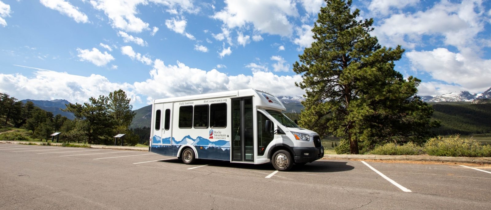 A white and blue shuttle bus is parked in an empty lot with trees, mountains, and a partly cloudy sky in the background.