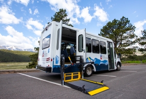White shuttle bus parked in a lot, with side doors open and a wheelchair lift extended onto the pavement; trees and mountains visible in the background.