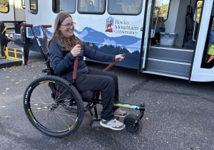 A woman in a wheelchair holds red straps near an accessible bus with a Rocky Mountain Conservancy logo in the background.