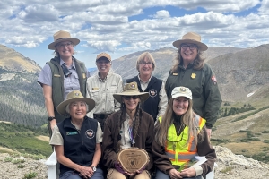 Seven women wearing park ranger uniforms and outdoor gear pose together on a mountain overlook, with one holding a wooden plaque. Mountains and cloudy sky are in the background.