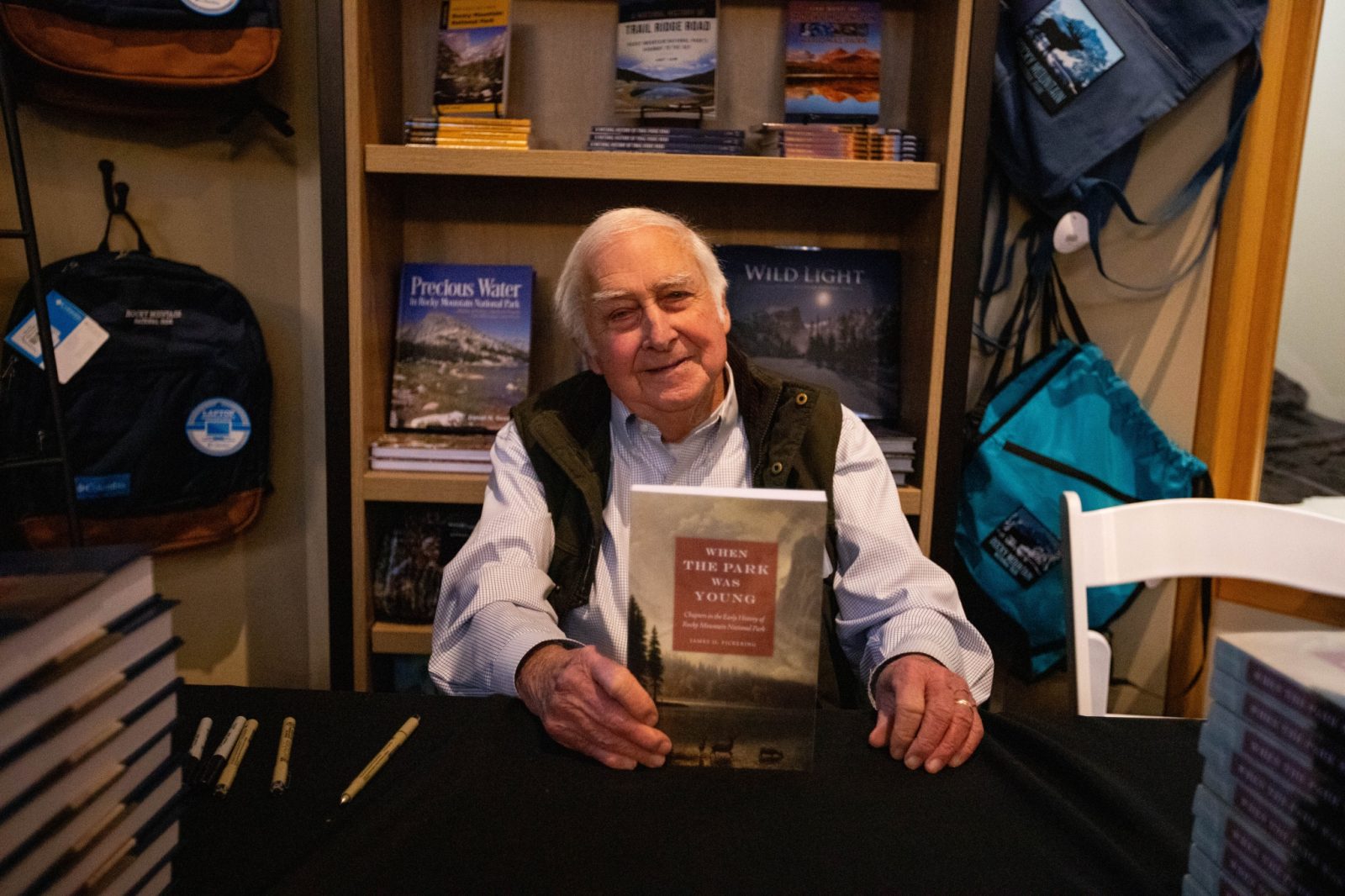 An elderly man sits at a table in a bookstore, smiling while holding up a copy of the book "When the Park Was Young." Stacks of books and pens are on the table.