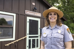 A park ranger in uniform stands in front of a brown building with a sign that reads "Volunteers in Parks.