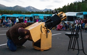 A person kneels to look through a large wooden telescope in a parking lot, with people, buses, and mountains visible in the background.