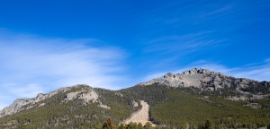 A mountain range with rocky peaks and dense forest under a clear blue sky. A visible rockslide or cleared path runs down one section of the slope.