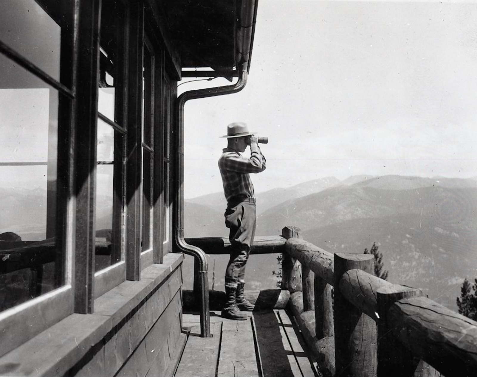 A person stands on a wooden lookout tower deck, wearing outdoor clothes and a hat, using binoculars to observe a mountainous landscape in the distance.