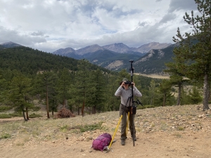 A person stands on a rocky hillside with surveying equipment and a pink backpack, surrounded by pine trees and mountains under a cloudy sky.