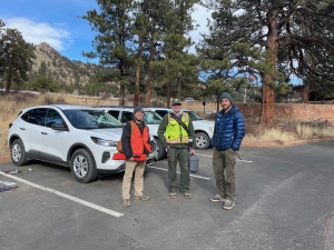 Three men wearing outdoor gear stand beside two white SUVs in a parking lot with trees and rocky hills in the background.