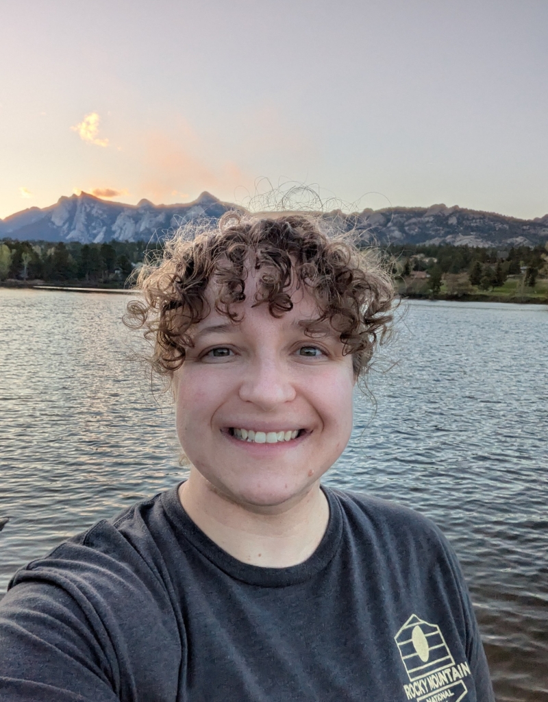 A person with curly hair smiles at the camera while standing by a lake, with mountains and trees visible in the background at sunset.
