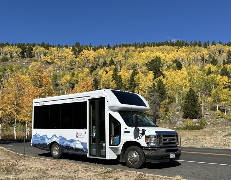 A white shuttle bus with mountain graphics is parked on the roadside, with a backdrop of trees showing fall colors and a clear blue sky.