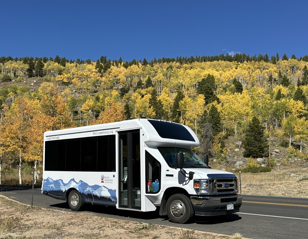A white shuttle bus with mountain graphics is parked on the roadside, with a backdrop of trees showing fall colors and a clear blue sky.